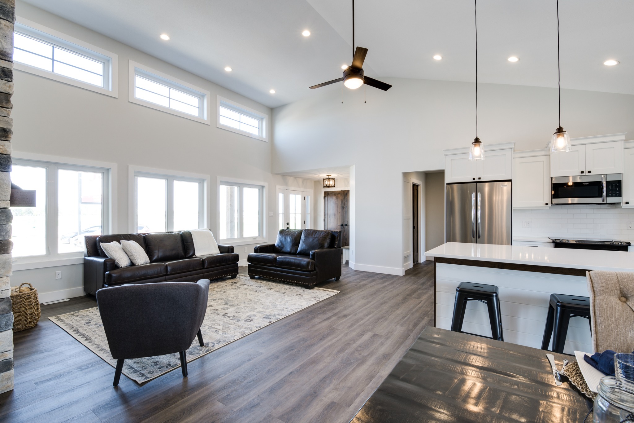 Spacious, modern open-plan living room and kitchen with high ceilings, pendant lights, dark wood floors, black leather sofas, and large windows.