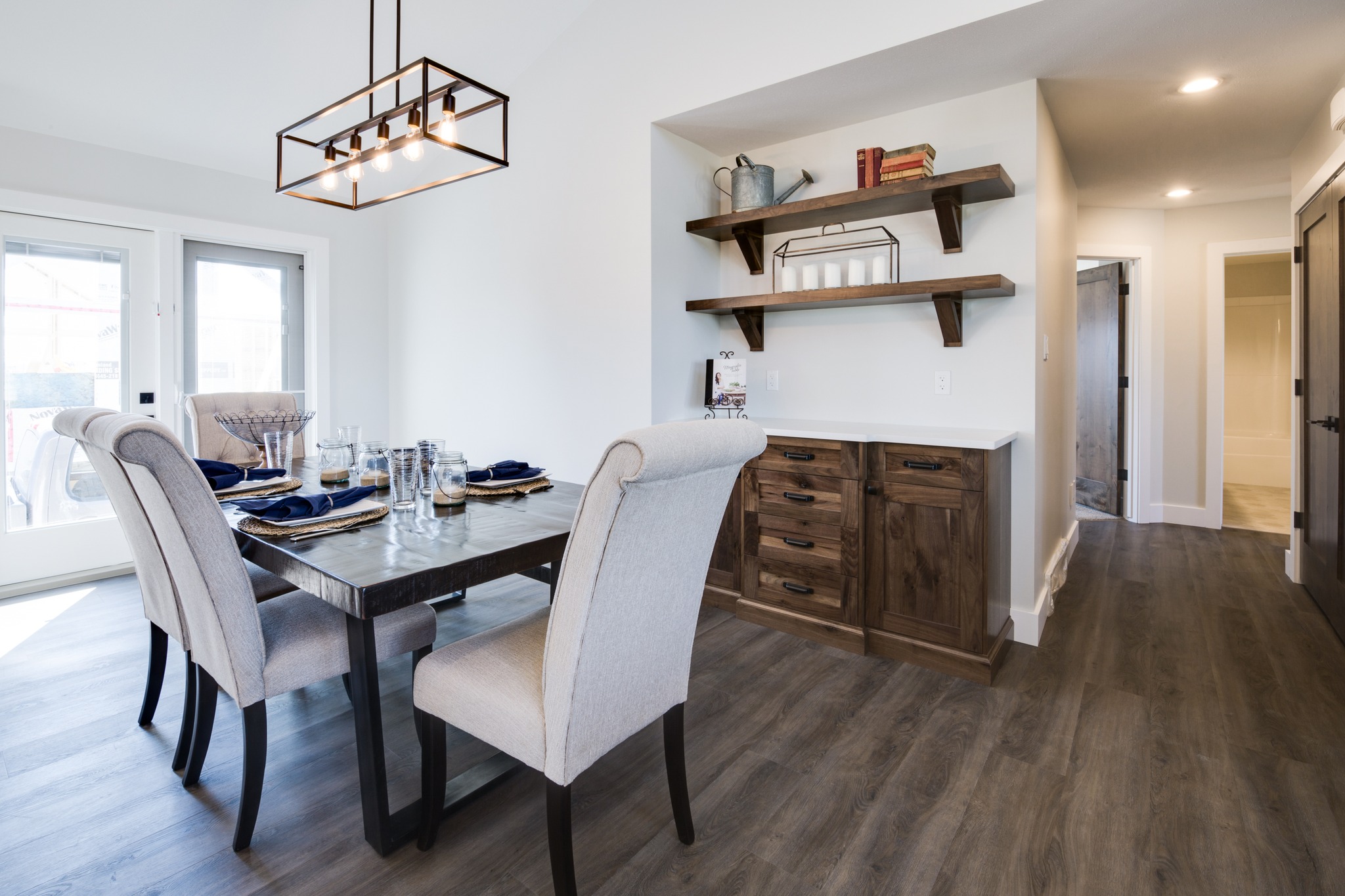 Modern dining room with light gray upholstered chairs around a dark wood table, set for four. Pendant lighting, wood flooring, shelving with décor. Bright, airy space.