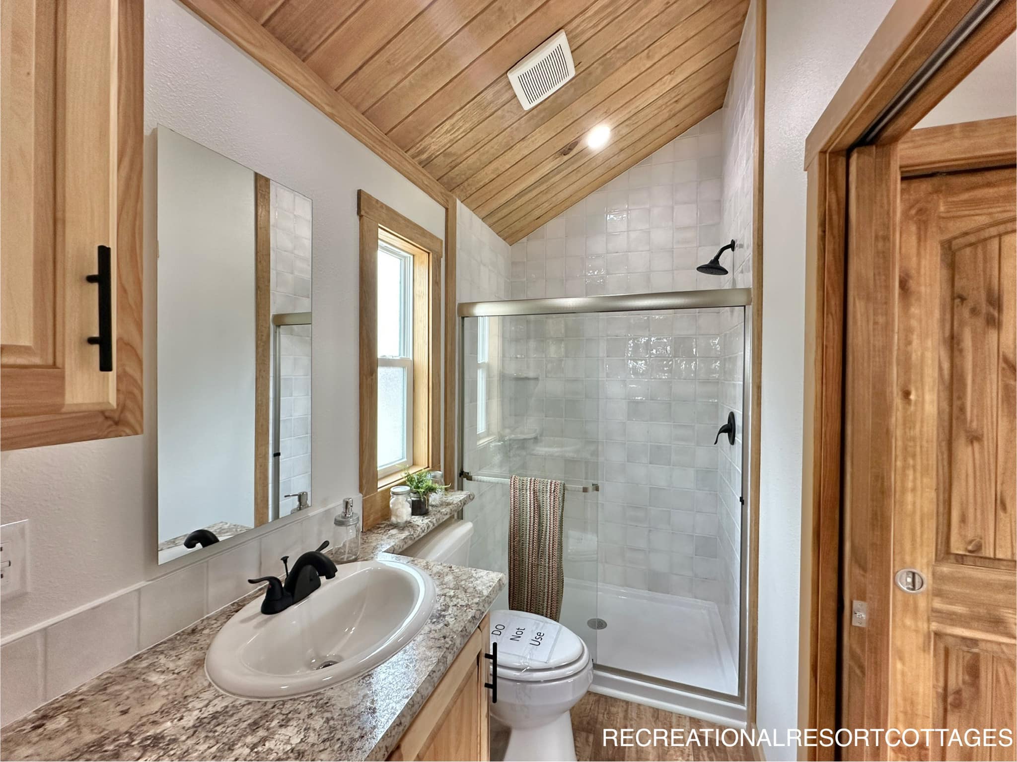 Cozy bathroom with wooden ceiling and door, featuring a glass-enclosed shower, white sink on a granite countertop, and a window for natural light.