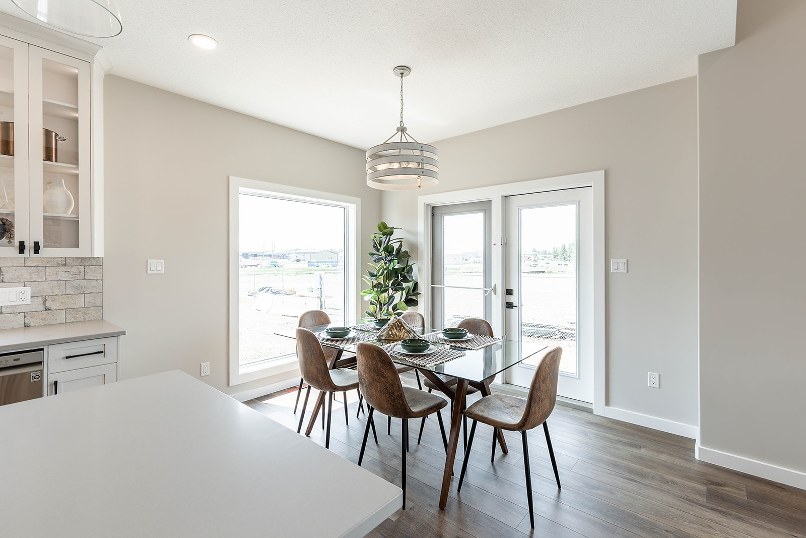 Modern dining area with a glass table set for six, surrounded by brown chairs. A potted plant stands by large windows and a door, enhancing brightness.