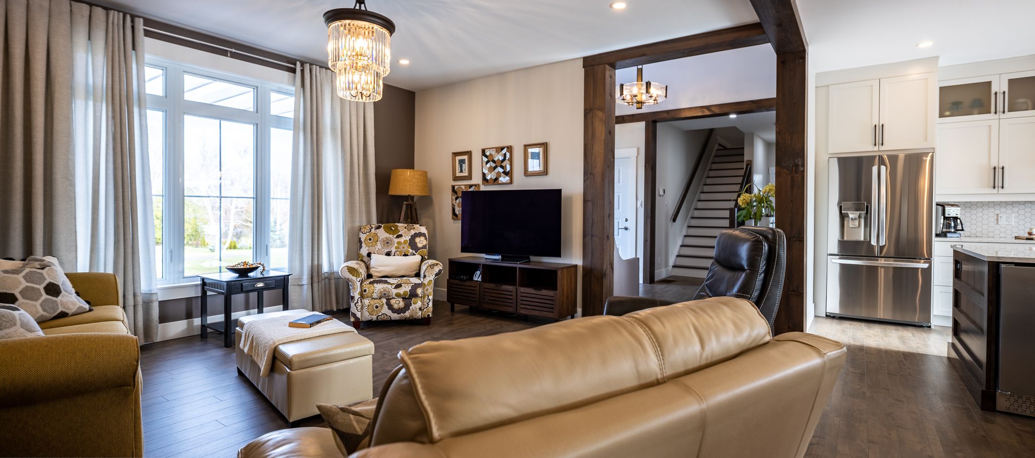 A cozy living room with elegant decor, featuring a floral armchair, leather sofas, large windows with curtains, a TV, and a kitchen in the background.