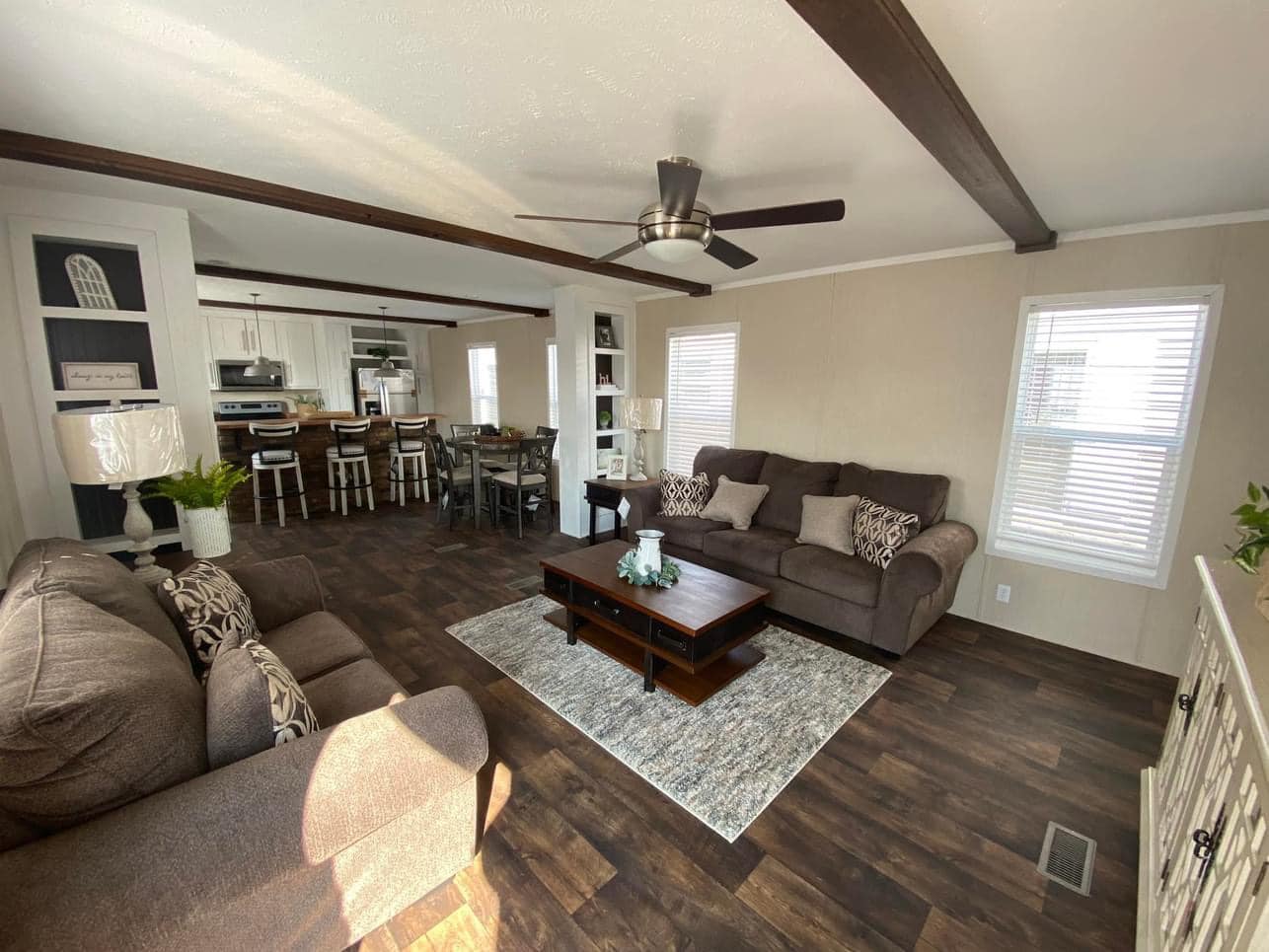 Spacious living room with brown sofas, patterned cushions, and a central wooden coffee table on a rug. Ceiling fan and exposed beams add a cozy feel.