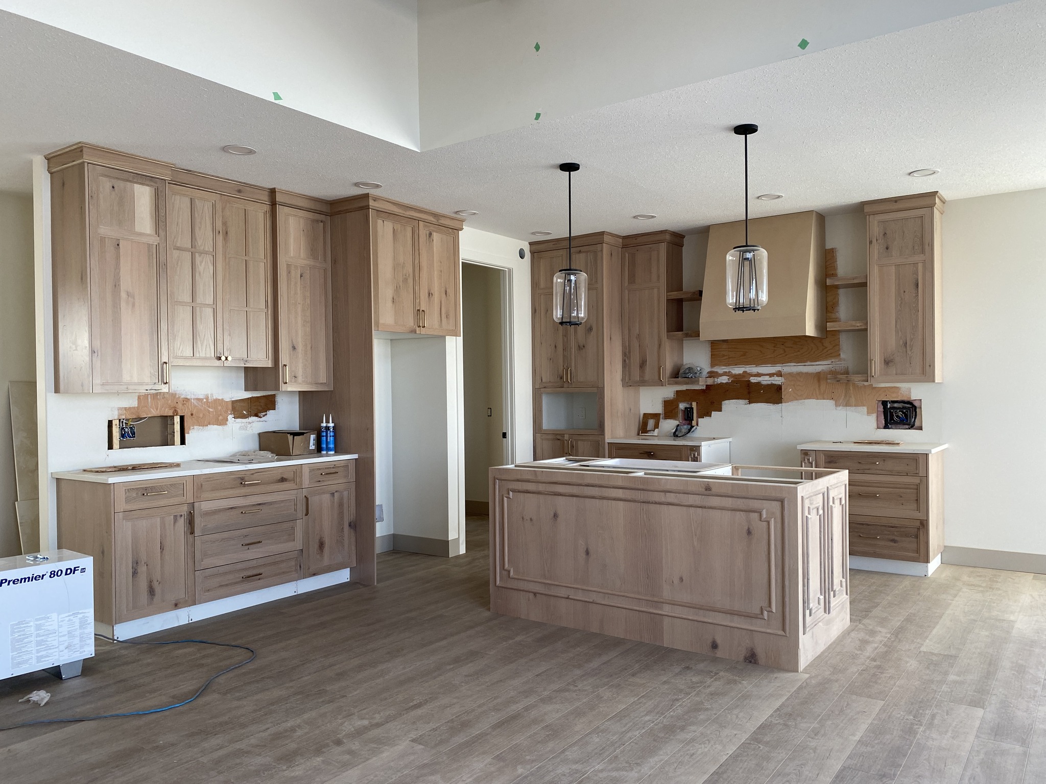 Modern kitchen under construction with natural wood cabinets and an island. Two pendant lights hang above, and the floor is light wood. Ambient lighting suggests a calm, welcoming atmosphere.