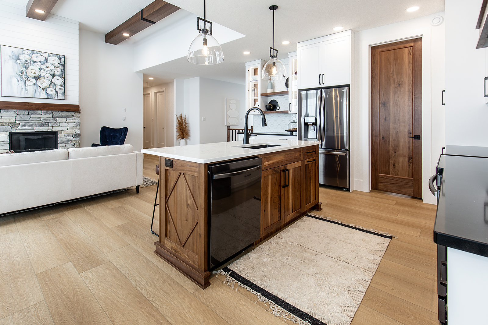 Modern kitchen with wood accents, white cabinetry, and a central island featuring a black dishwasher. The space is open, leading to a cozy living area with a stone fireplace and large artwork. The atmosphere is warm and inviting.