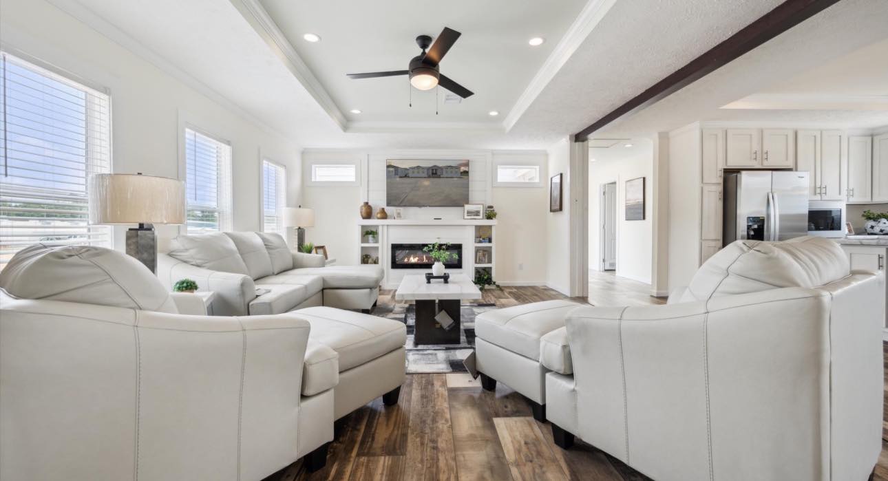 Modern living room with white walls and furniture, featuring a ceiling fan, recessed lighting, and a cozy fireplace. Large windows illuminate the space.