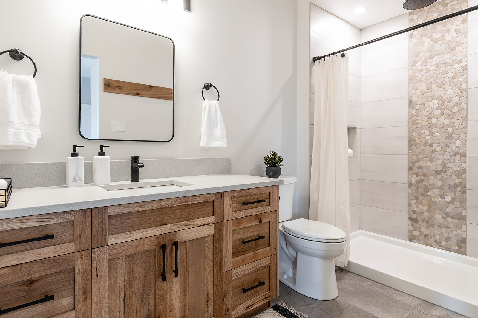 Modern bathroom featuring a wooden vanity with black hardware, a rectangular mirror, and twin towel holders. The shower has a neutral tile accent. Bright and minimalistic.