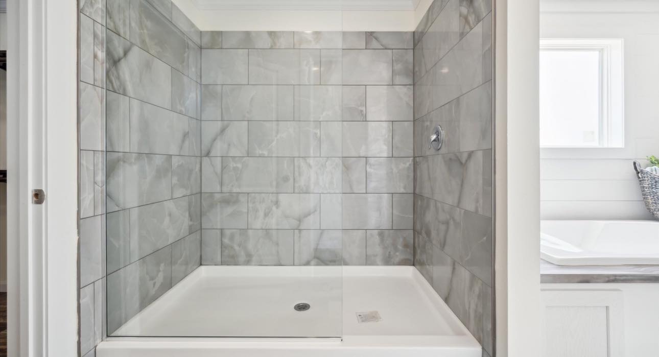 A modern shower with elegant gray marble tiles and a glass door, conveying a sleek, clean look. Sunlight filters in from a nearby window, adding warmth.