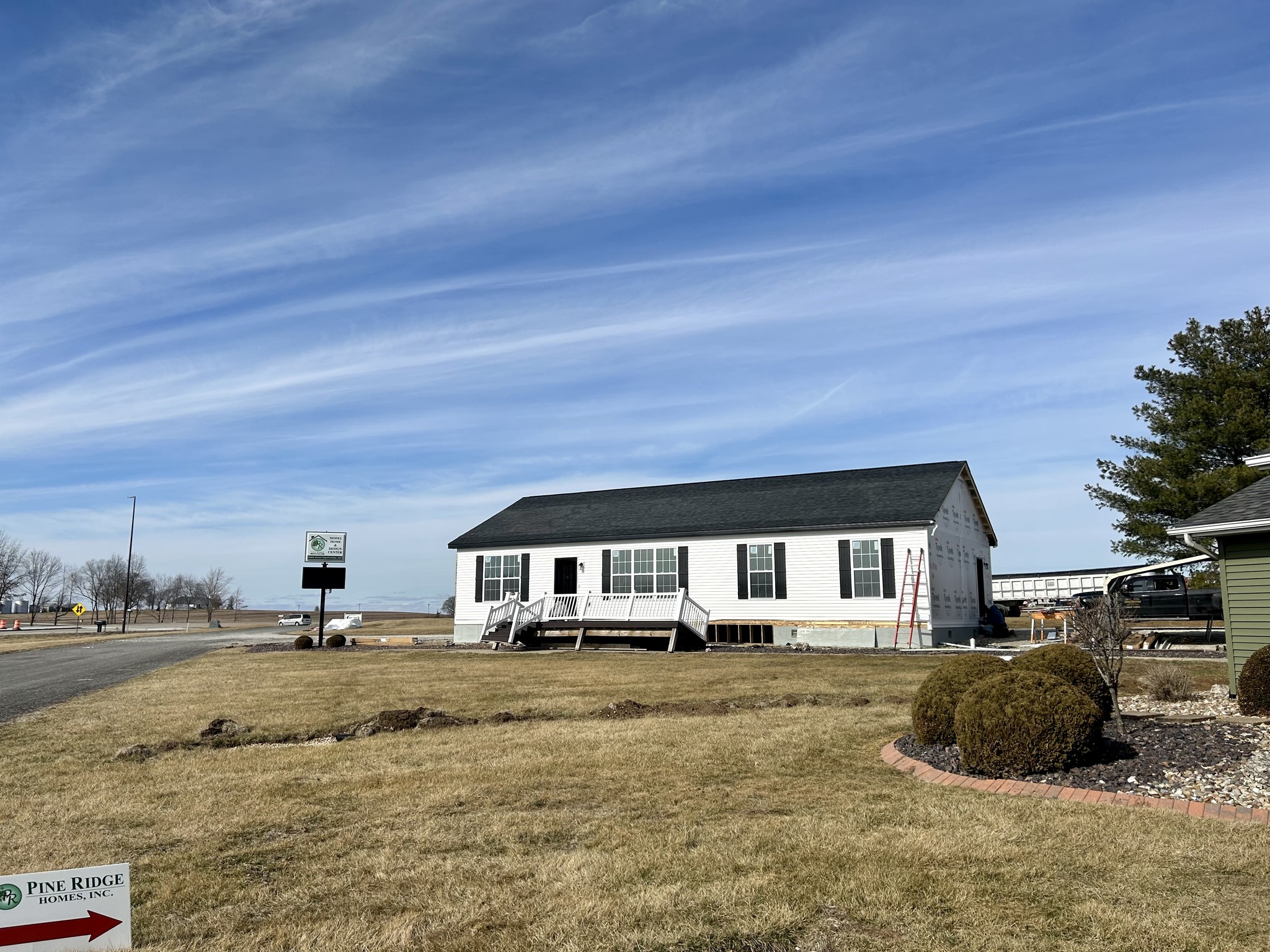 A white modular home with a dark roof is on a grassy lot under a clear blue sky. A red ladder leans against it. Nearby, bushes and a sign read "Pine Ridge Homes, Inc."