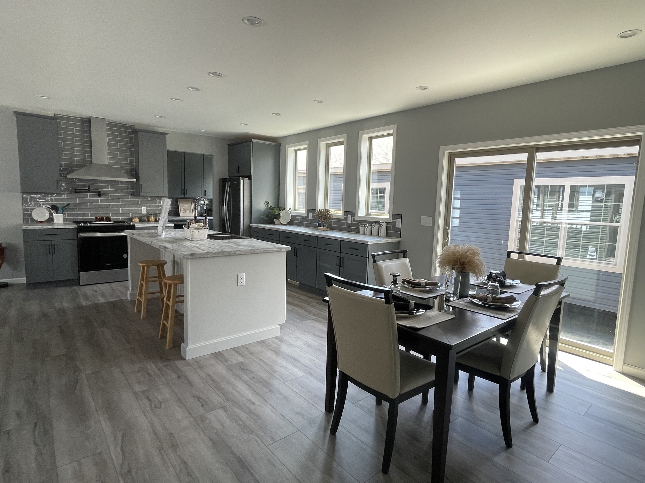 Modern kitchen and dining area with gray cabinets, marble island, stainless appliances, and a set dining table by large windows, creating a bright, airy feel.