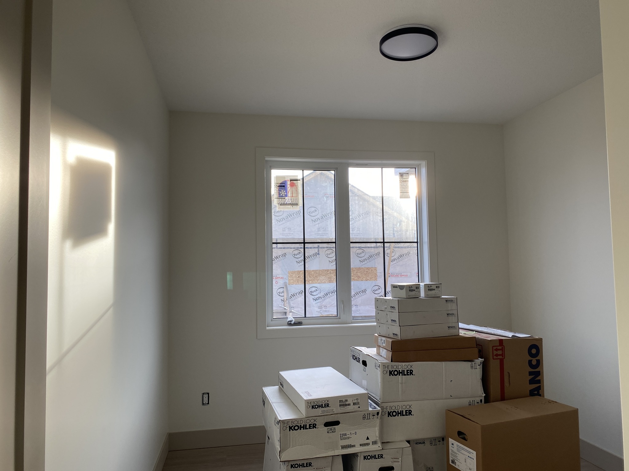 Sunlit room with a window and a pile of stacked boxes labeled "Kohler" near the corner. Walls are bare, and a round ceiling light is visible.