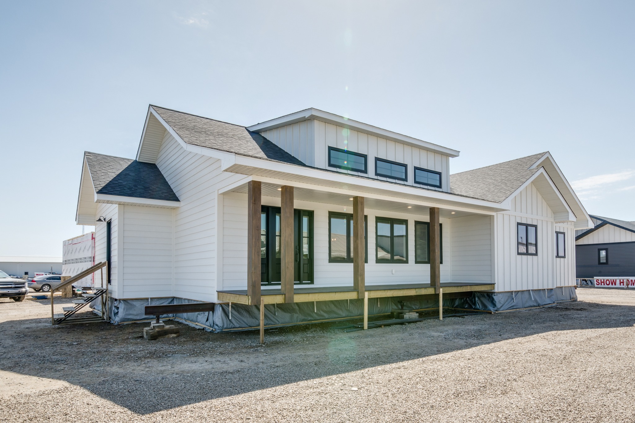 A modern, white modular home with a dark roof and large windows sits on a gravel lot under a clear blue sky, evoking a sense of simplicity and neatness.