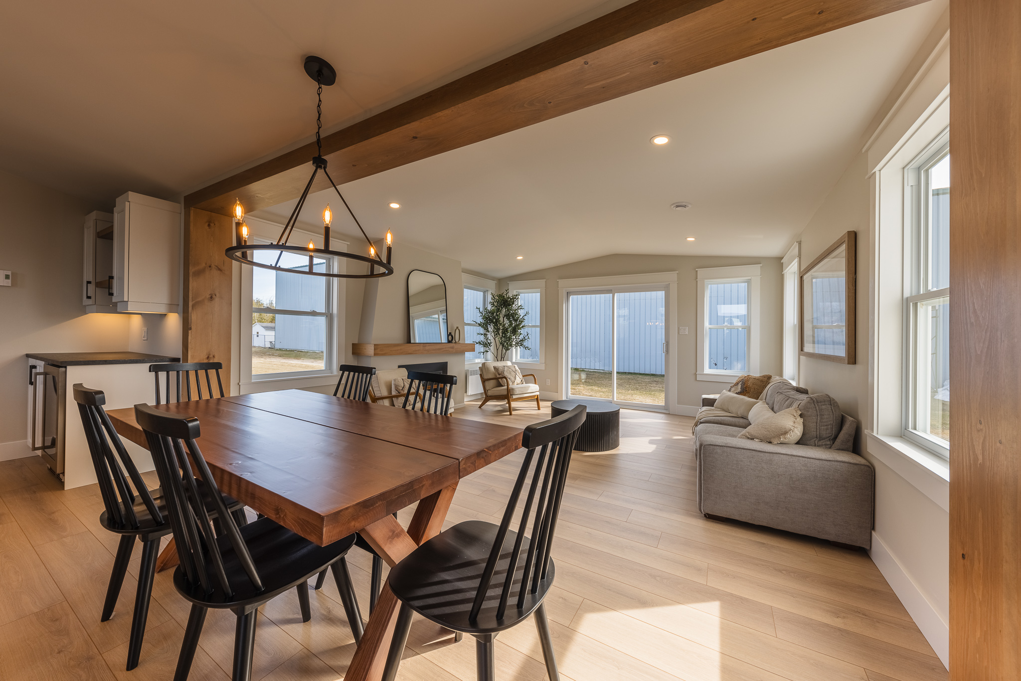 Modern open-plan living and dining area with wooden floors. Features a wooden table with black chairs, a cozy gray sofa, large windows, and warm lighting.