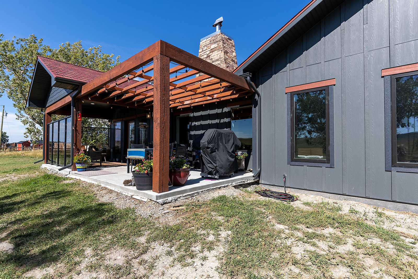 Modern house exterior featuring a shaded patio with wooden pergola, a stone chimney, and plants. The building has gray walls and large windows.