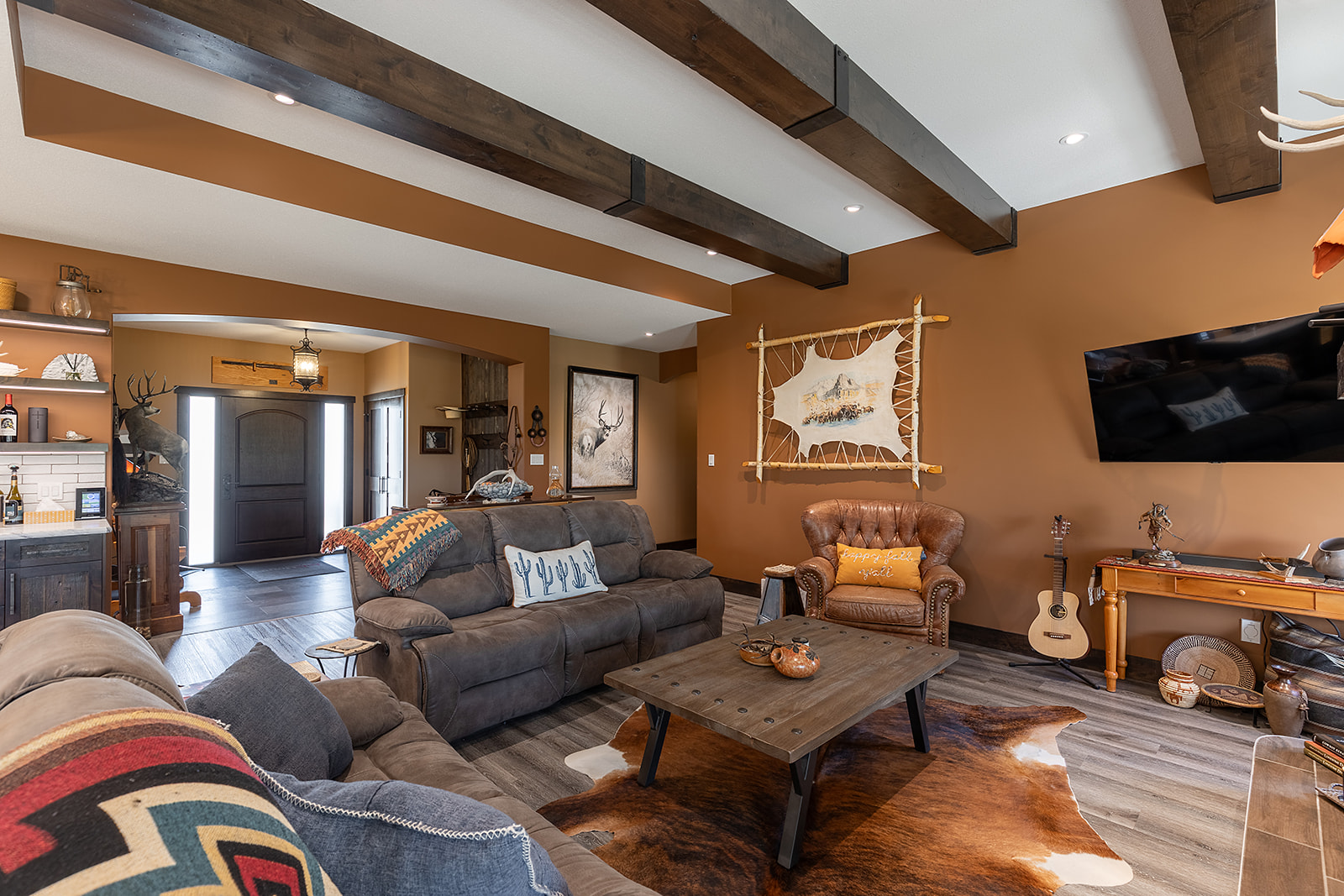Cozy living room with rustic decor and warm tones. Features a leather armchair, textured couches, wood beams, a wall-mounted TV, and a guitar by the wall.