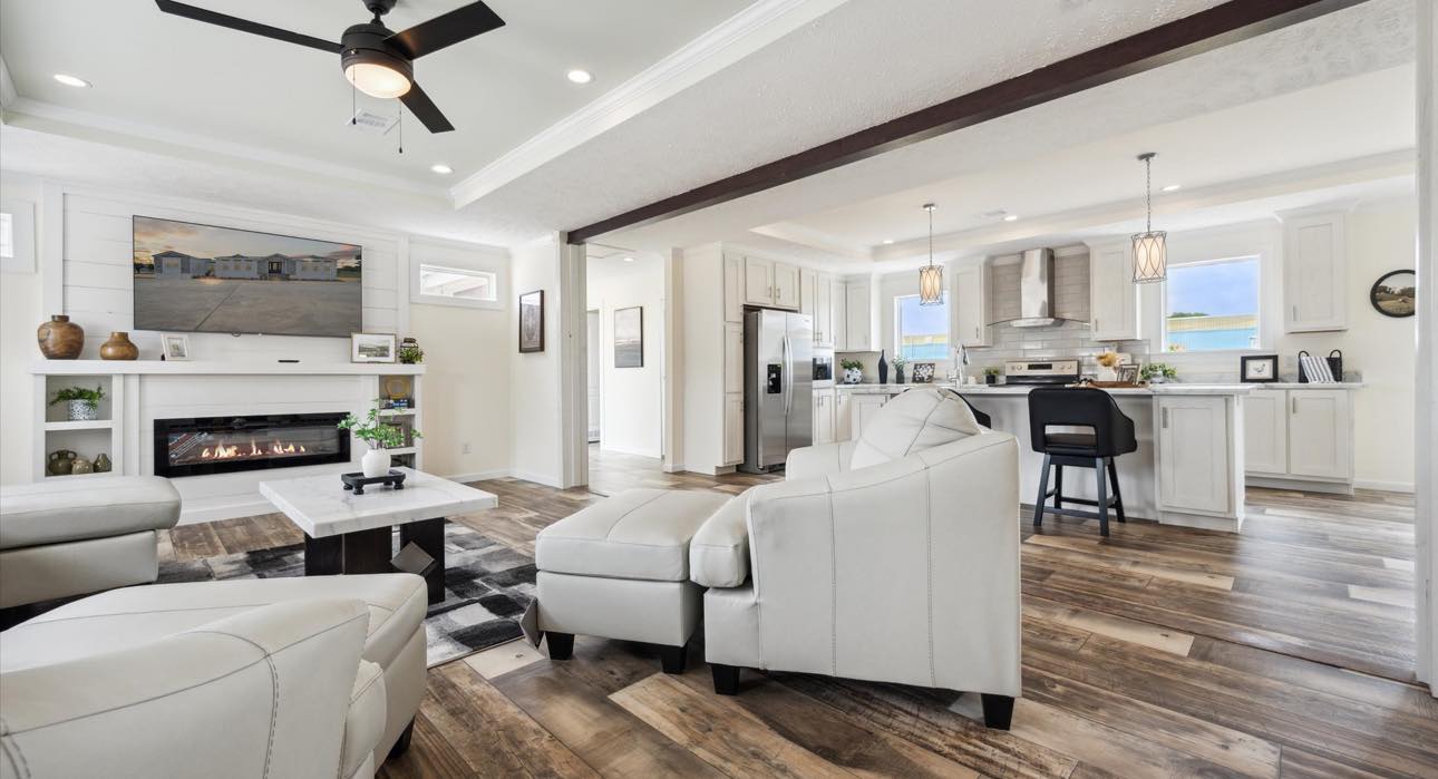Spacious modern living room and kitchen with white furniture, wood flooring, ceiling fan, and cozy fireplace, creating a warm, inviting atmosphere.
