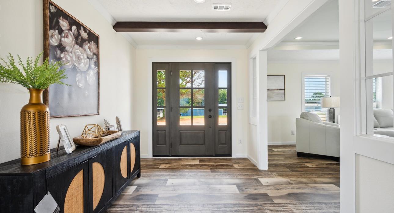 A bright entryway with dark wooden double doors, wooden floor, and a modern console table on the left. A comfy living room is visible to the right.