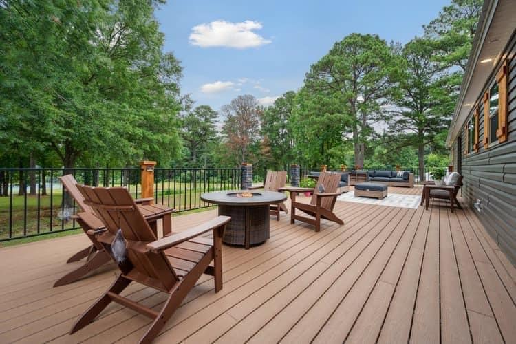 Spacious wooden deck with Adirondack chairs, a fire pit, and distant outdoor seating. Surrounded by lush trees under a clear blue sky, creating a serene ambiance.