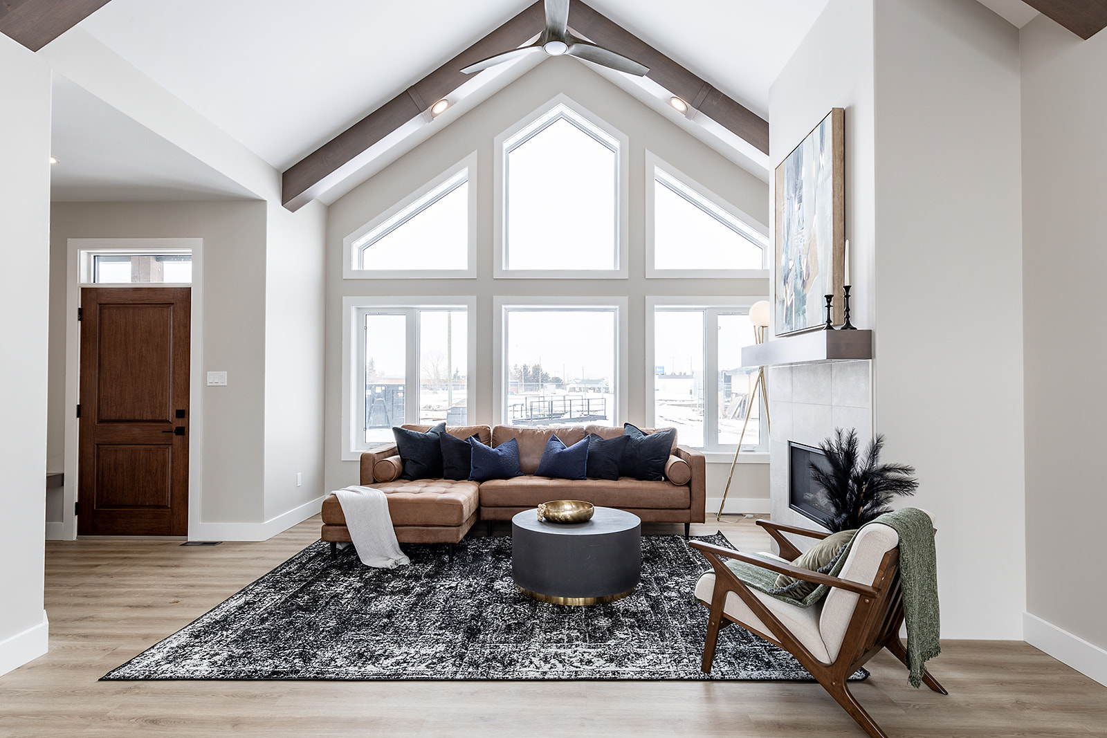 A modern living room with a vaulted ceiling, large windows, tan sectional sofa, black and white rug, fireplace, and a wooden chair, conveying warmth.