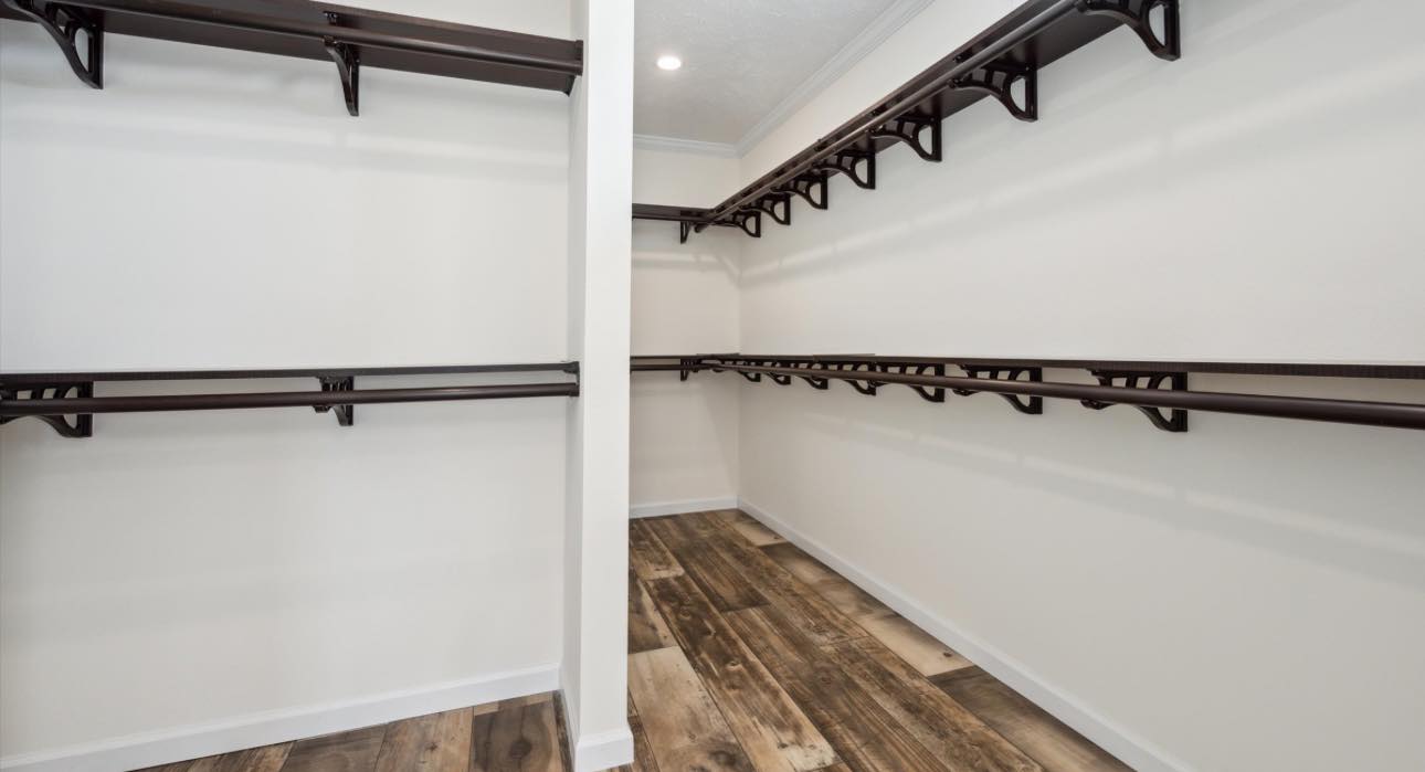Spacious, empty walk-in closet with white walls and wooden floors. Dark brown wooden shelves and rods line the sides. Bright and clean atmosphere.