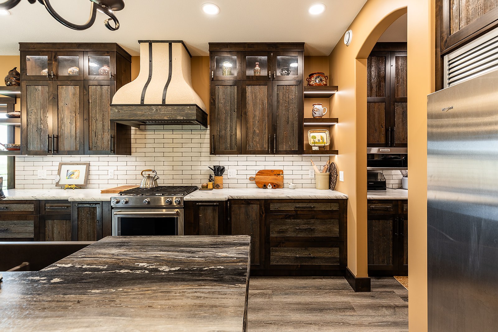 Warm-toned kitchen with rustic wood cabinets, sleek stainless steel appliances, and white brick backsplash. Marble countertops hold a teakettle and utensils.