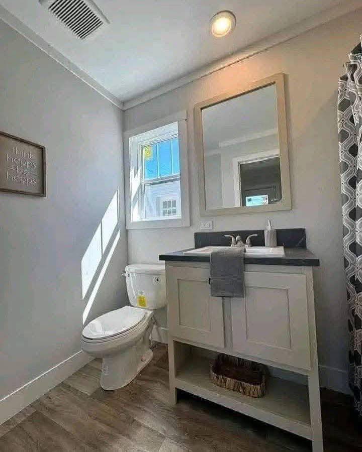 A small bathroom with a white toilet beside a vanity with a mirror. Sunlight from a window casts shadows. There's a "Think Happy Be Happy" sign on the wall.