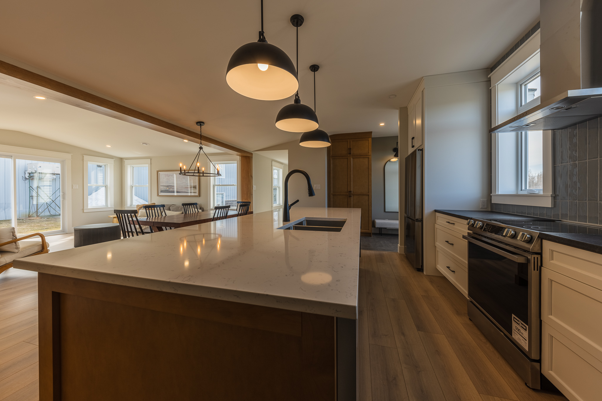 Modern kitchen with a large island, sleek black fixtures, and pendant lights. The open space extends to a dining area with a chandelier, creating a warm and inviting atmosphere.