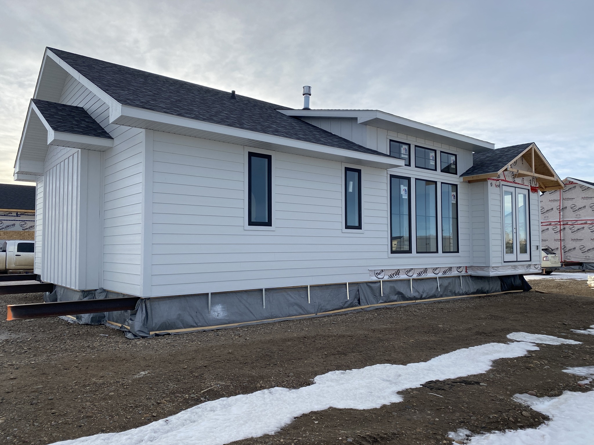 A modern, white house under construction, showcasing large windows and a dark roof. The ground is bare soil with patches of snow, under a cloudy sky.