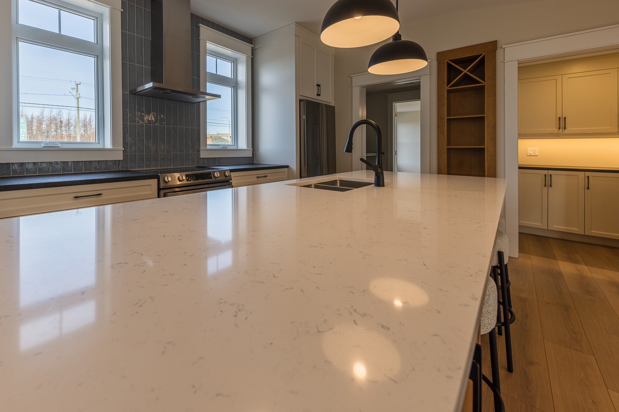 Modern kitchen with a large marble island, black faucet, and pendant lighting. Bright windows illuminate wooden flooring and cabinets, creating a sleek and inviting atmosphere.