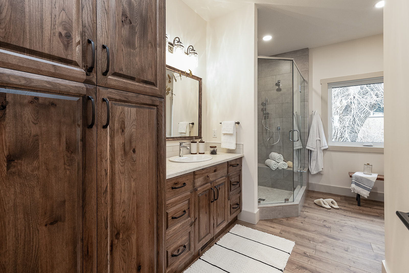 Stylish bathroom with warm wood cabinets, a large mirror, and modern lighting above a double sink. Glass shower, cozy ambiance with natural light.