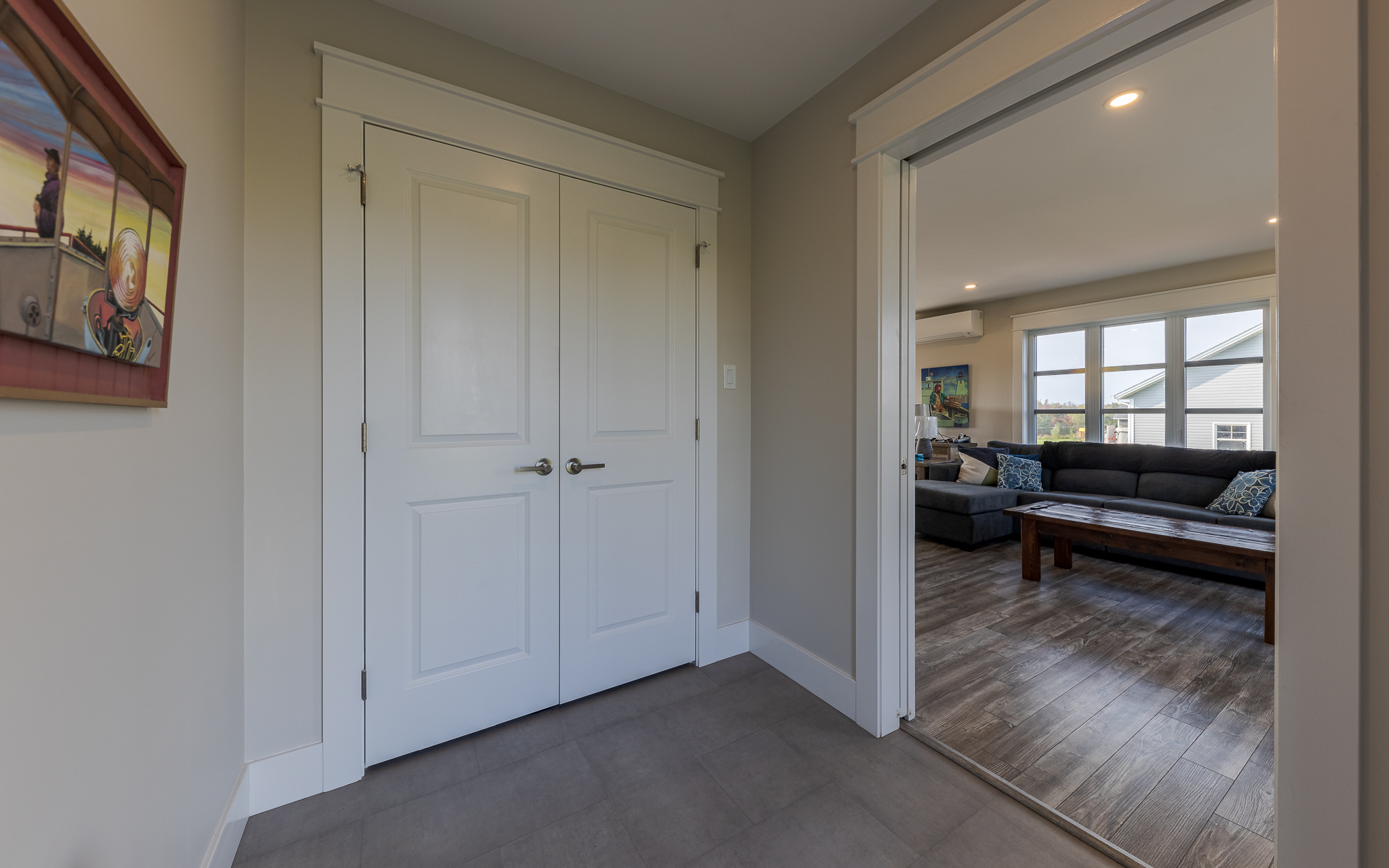 Hallway with gray tile flooring and a closed double door to the left. On the right, a doorway opens to a living room with a dark couch and large windows.