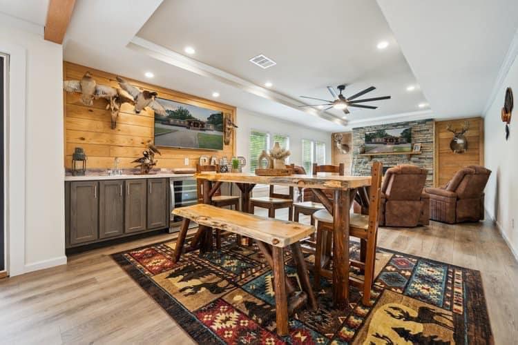 Rustic dining room with wooden table and benches on a patterned rug. Mounted deer heads on wood-paneled wall, modern ceiling fan, cozy recliners by a stone wall.