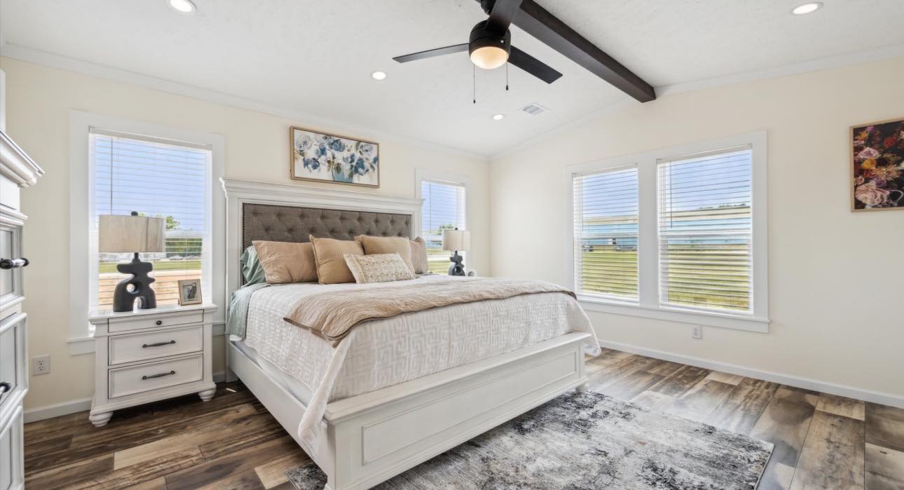 Cozy bedroom with a large bed, neutral tones, and decorative pillows. Sunlit windows, a ceiling fan, and hardwood floors create a warm, inviting atmosphere.