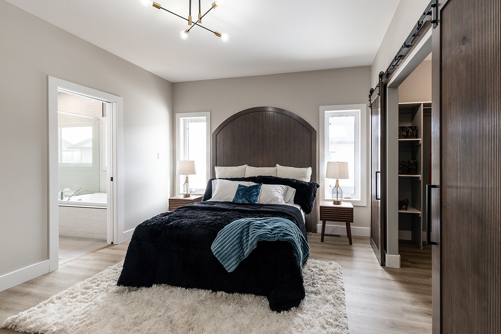 Modern bedroom with a dark wood headboard and plush bedding. Two side tables with lamps, a fluffy white rug, and a visible bathroom. Cozy and inviting.
