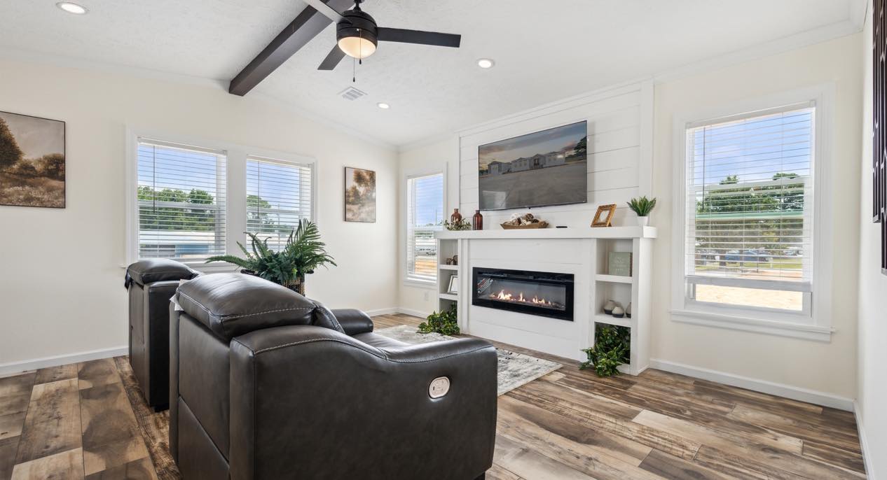 A cozy living room features dark leather recliners facing a modern electric fireplace. Large windows provide natural light, enhancing the warm, inviting atmosphere.