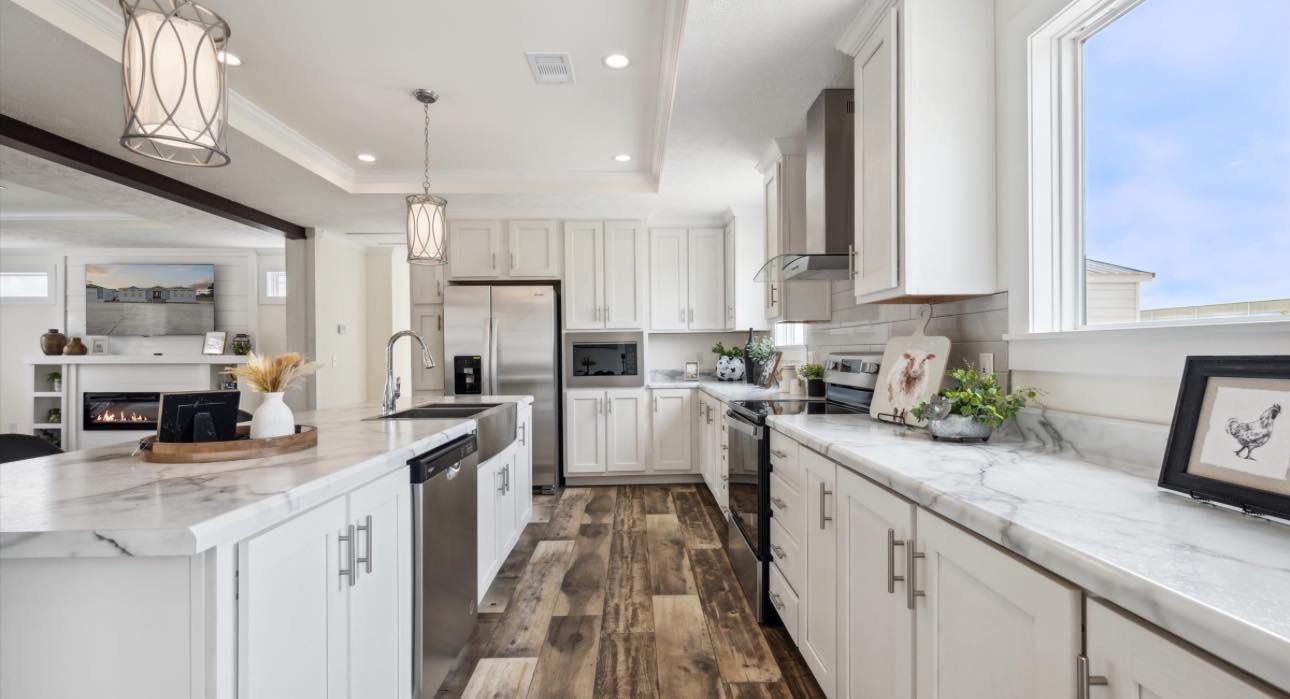 Bright, modern kitchen with white cabinets, marble countertops, and wood-style flooring. Large windows, pendant lights, and greenery add a fresh touch.