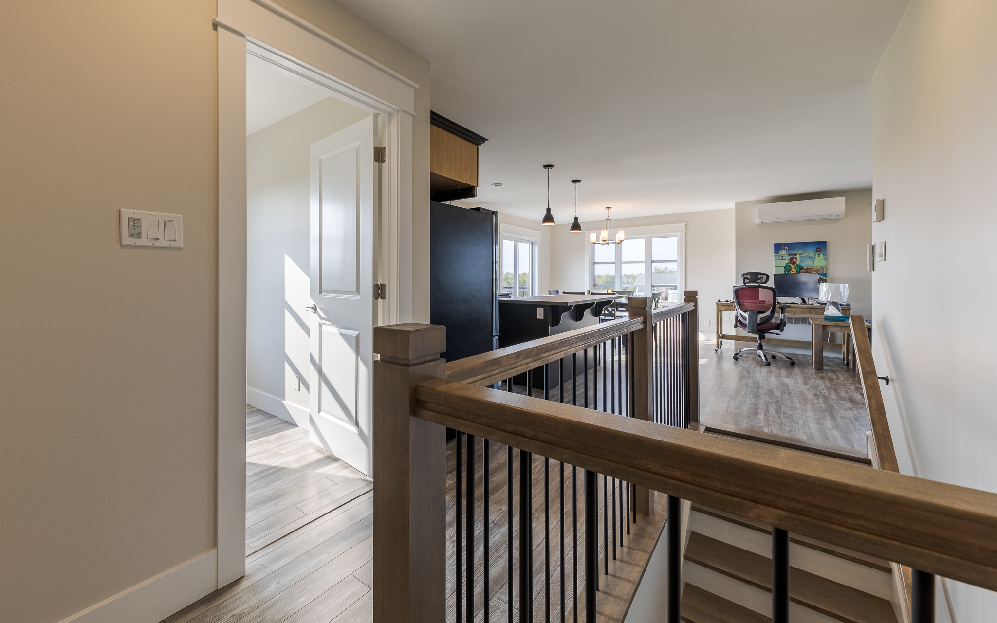 A modern, sunlit home interior with beige walls. Features include a staircase with wooden railings, a distant office area, and pendant lights. Calm ambiance.