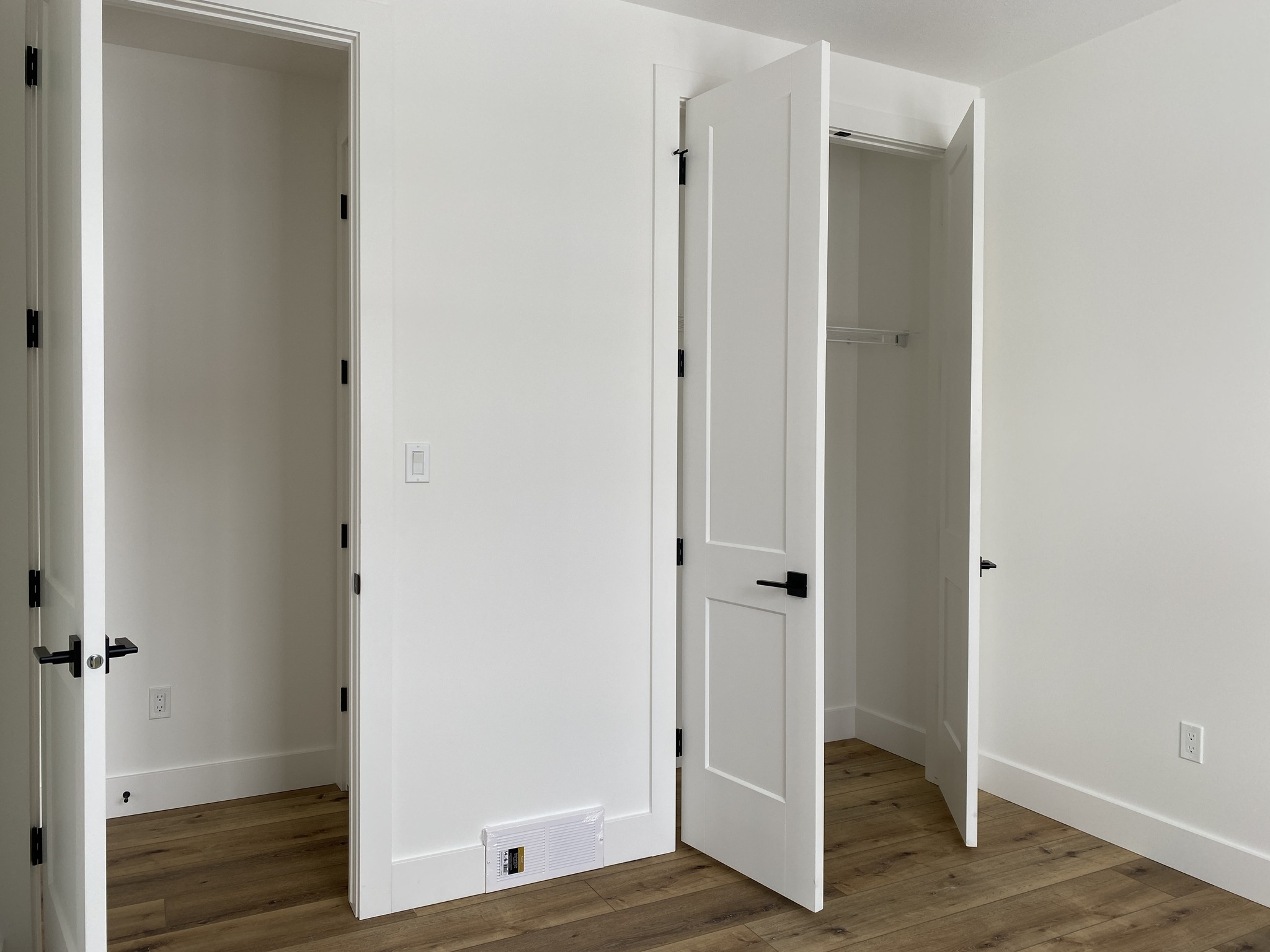 White room with light wood flooring, featuring two open closets with simple white doors and black handles. The space feels clean and minimalist.