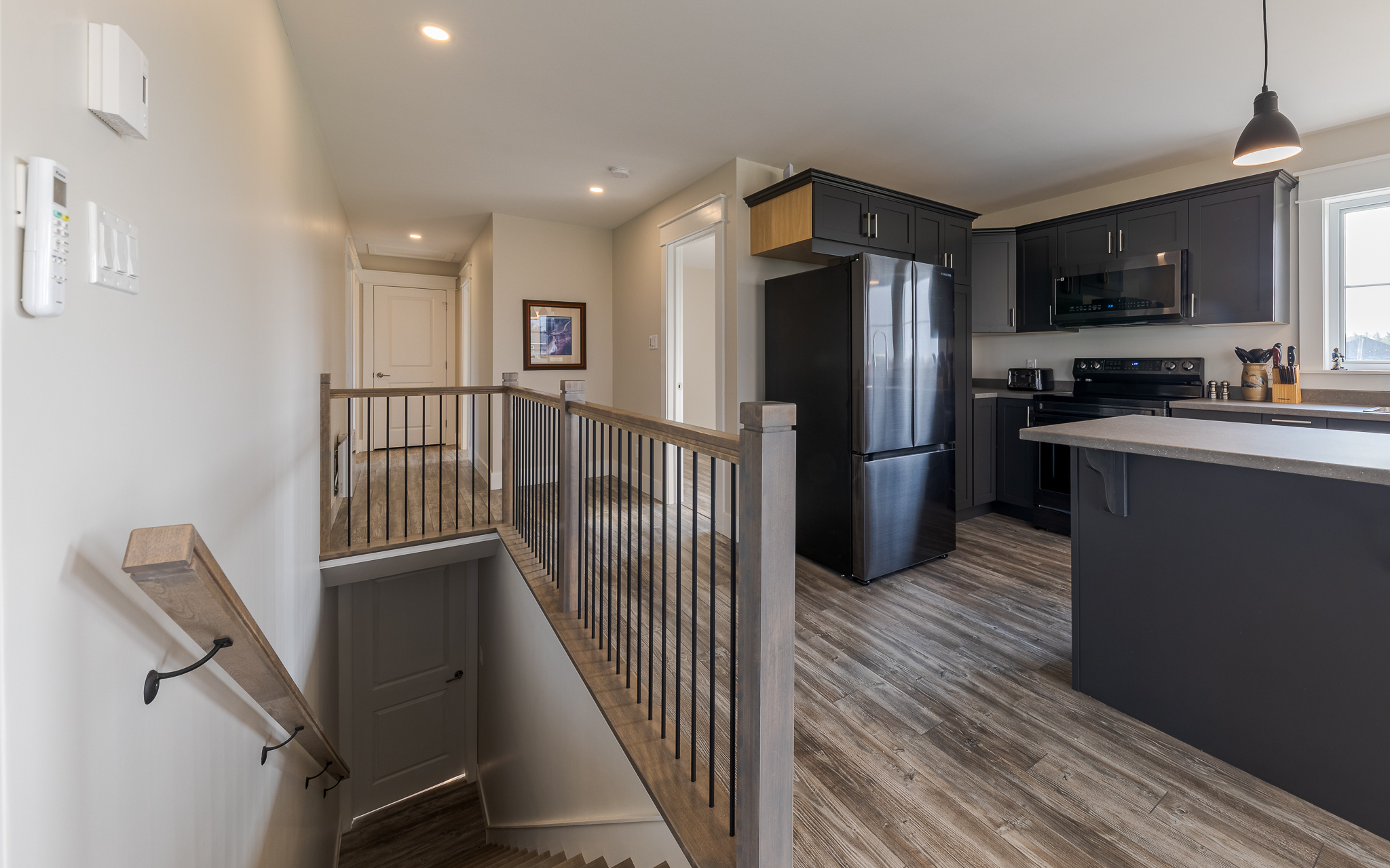 Modern kitchen and hallway with wood flooring. Dark cabinets and appliances, bright lighting, and an open staircase. Clean, welcoming atmosphere.