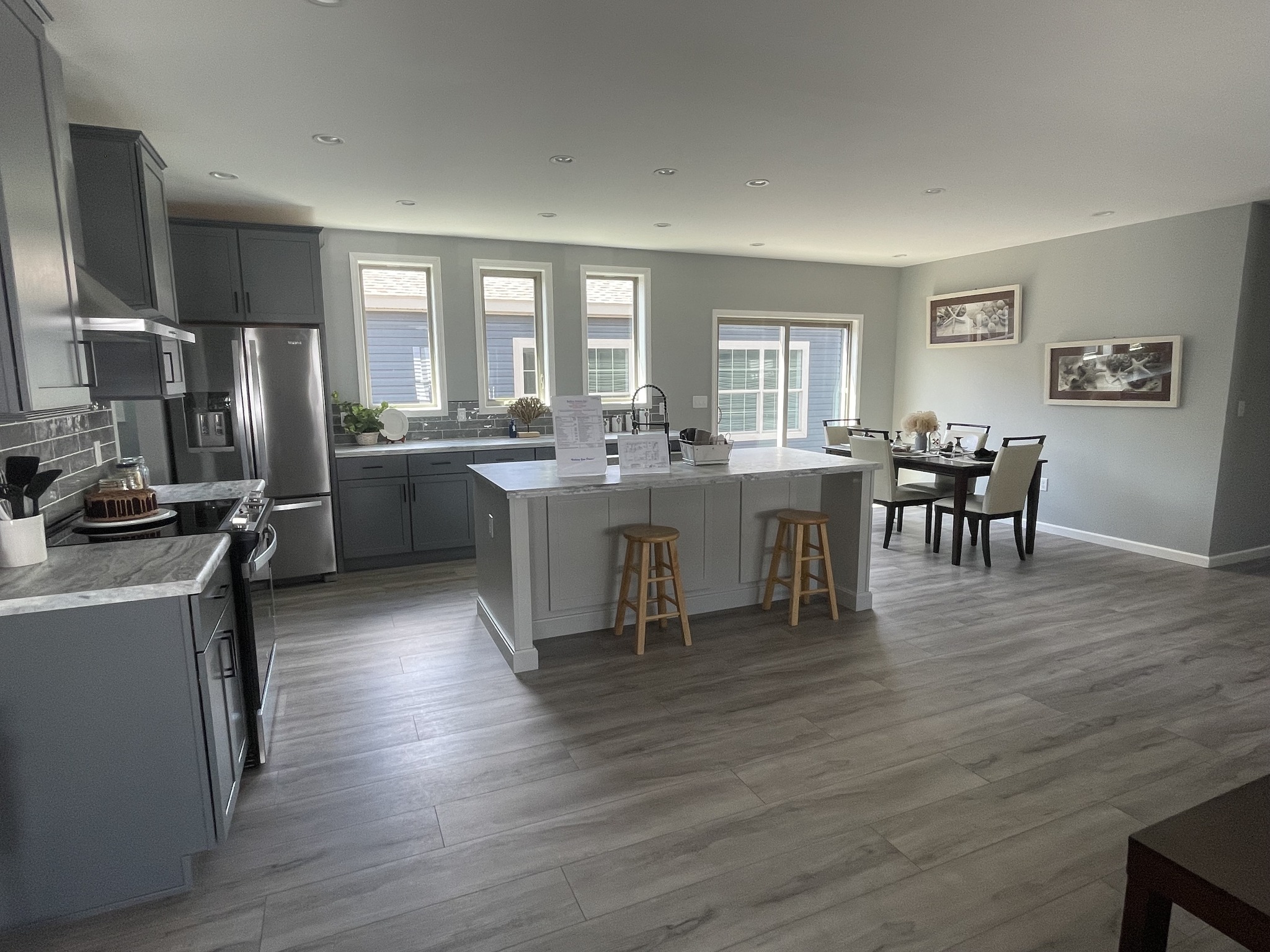 Spacious modern kitchen with gray cabinets, island with stools, stainless steel appliances, and adjacent dining area. Natural light from large windows.