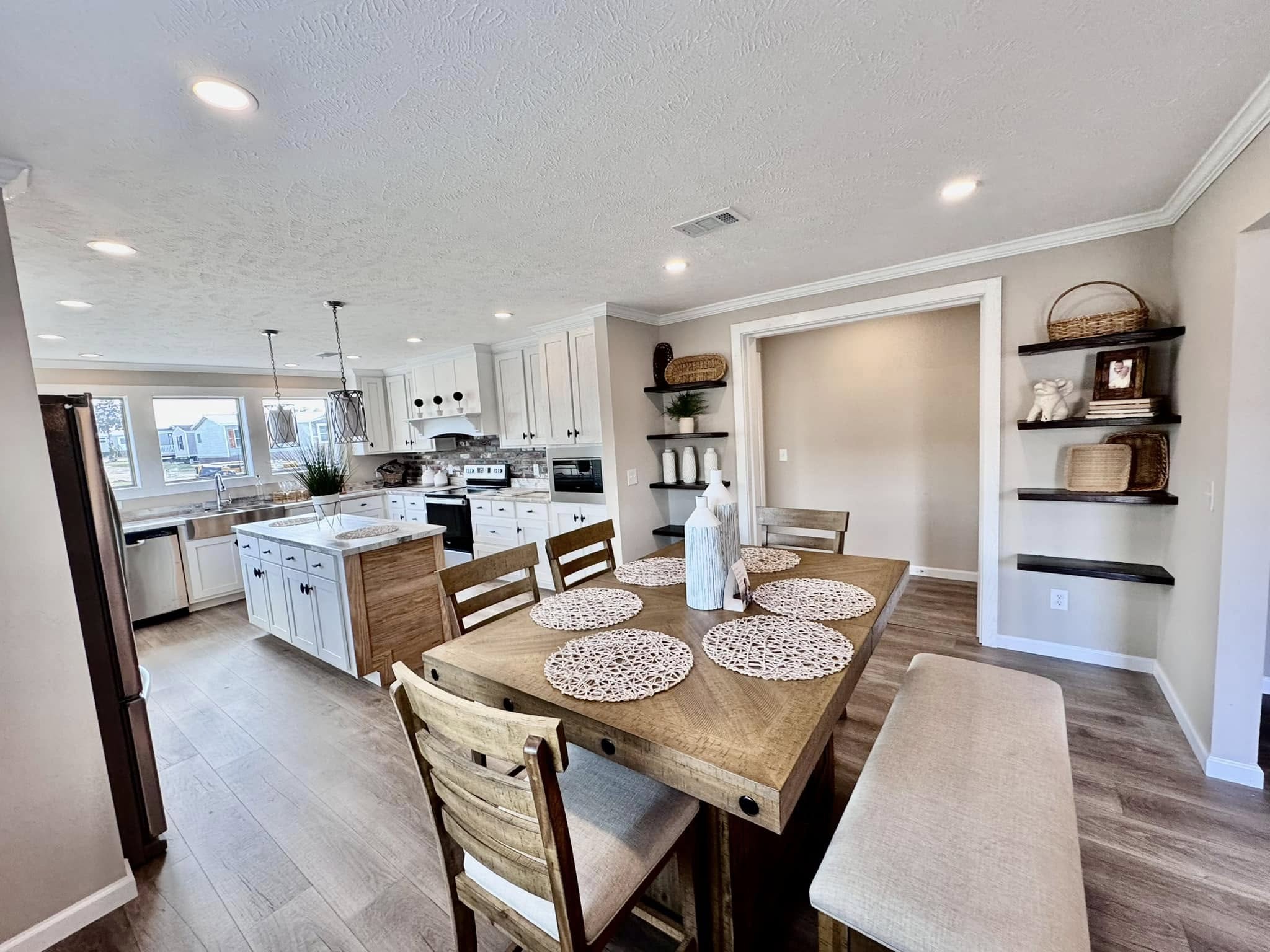 Spacious kitchen with wooden dining table, chairs, and bench, set with woven placemats. White cabinets, stainless steel appliances, and open shelves add a modern touch.