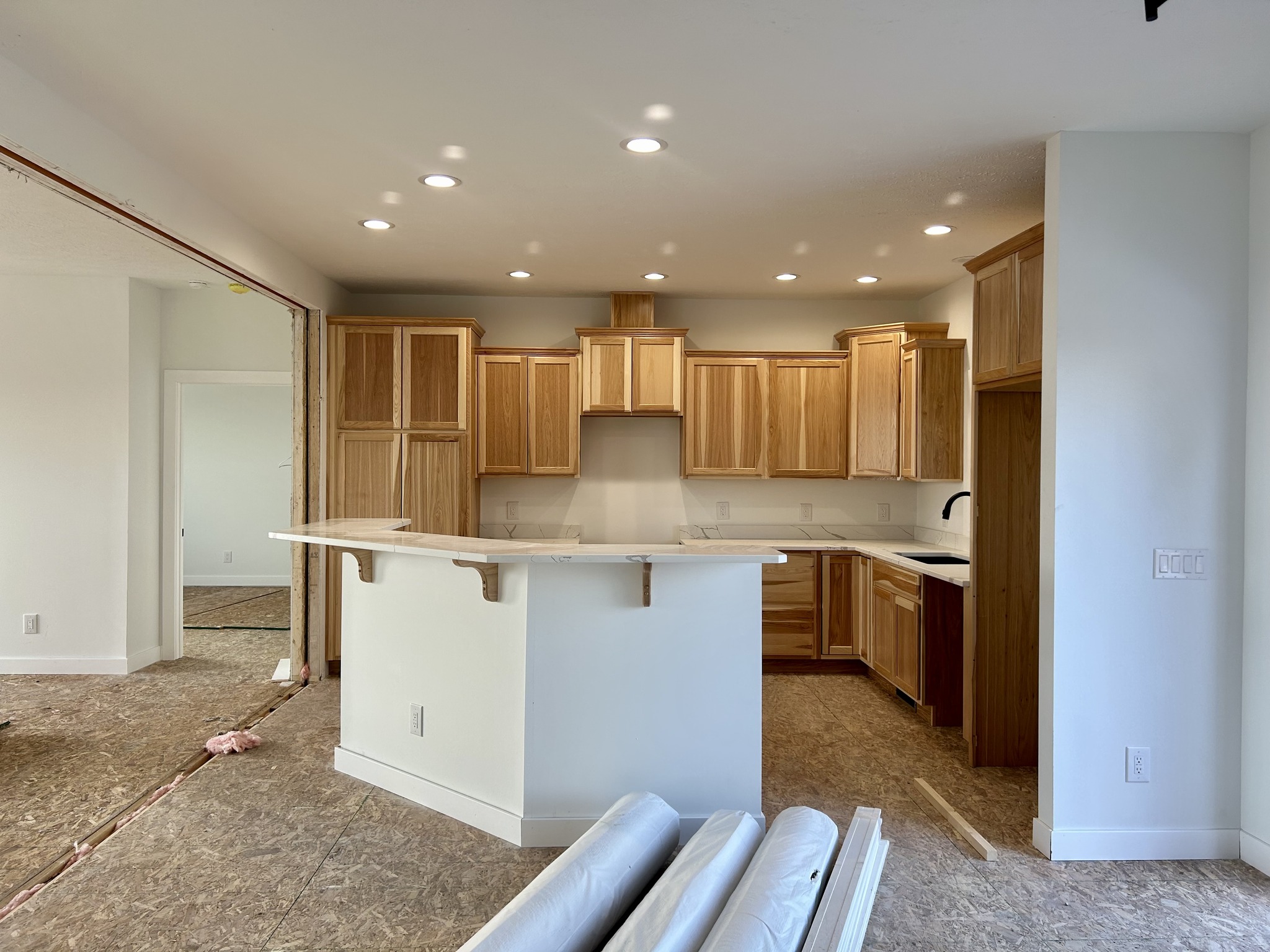 Spacious kitchen with light wood cabinets, white countertops, and a black faucet. Recessed lighting creates a warm, inviting ambiance. Construction materials visible.