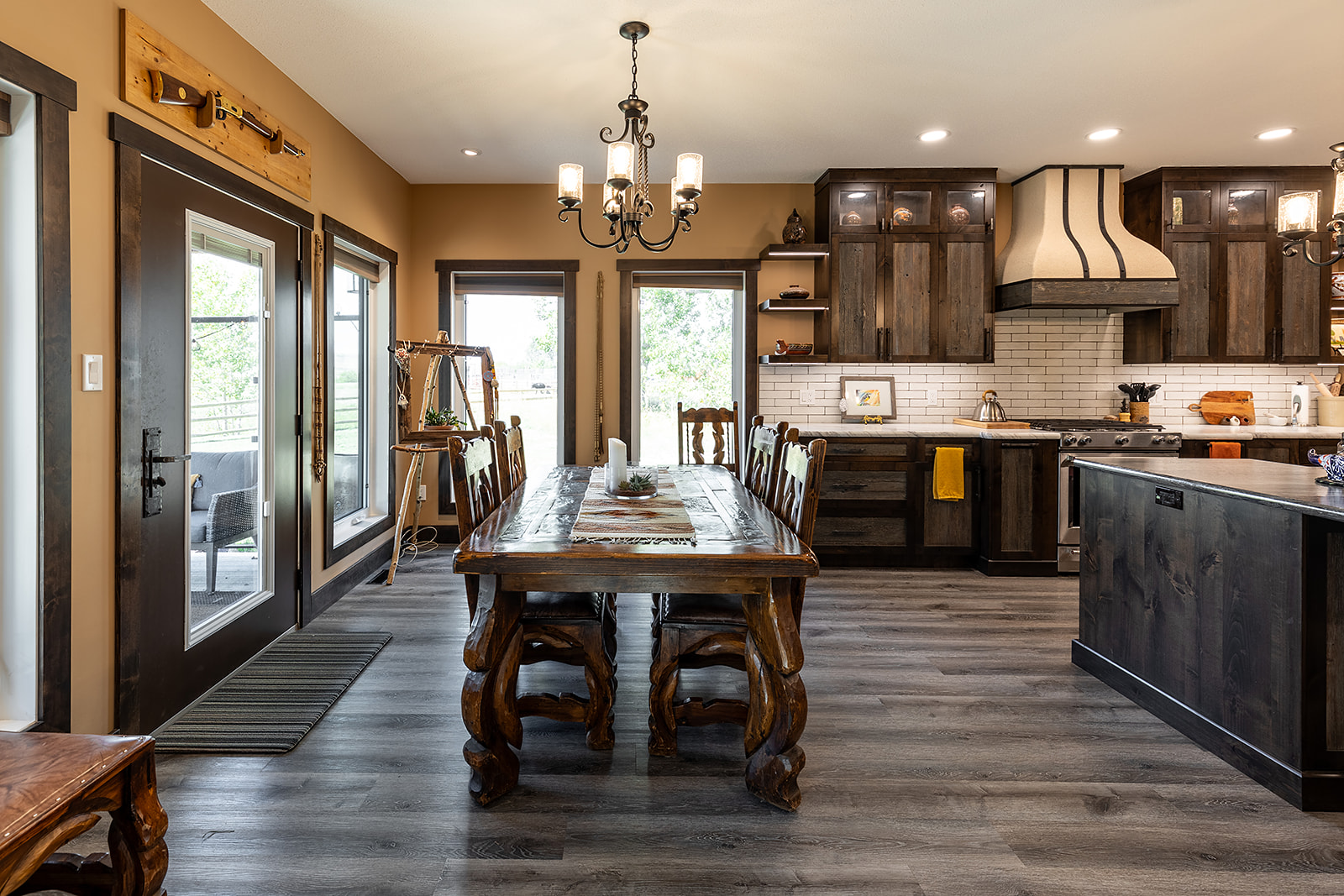 Warm, rustic dining and kitchen area with dark wood furniture, a large wooden table, and ornate chairs. Modern light fixtures and natural light create a cozy, inviting atmosphere.