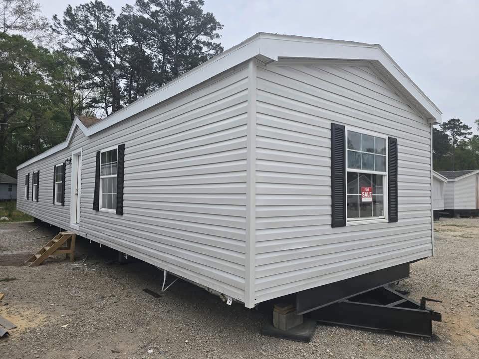 White mobile home with black shutters and a "For Sale" sign in the window, set on a gravel lot. Surrounded by trees under a cloudy sky.
