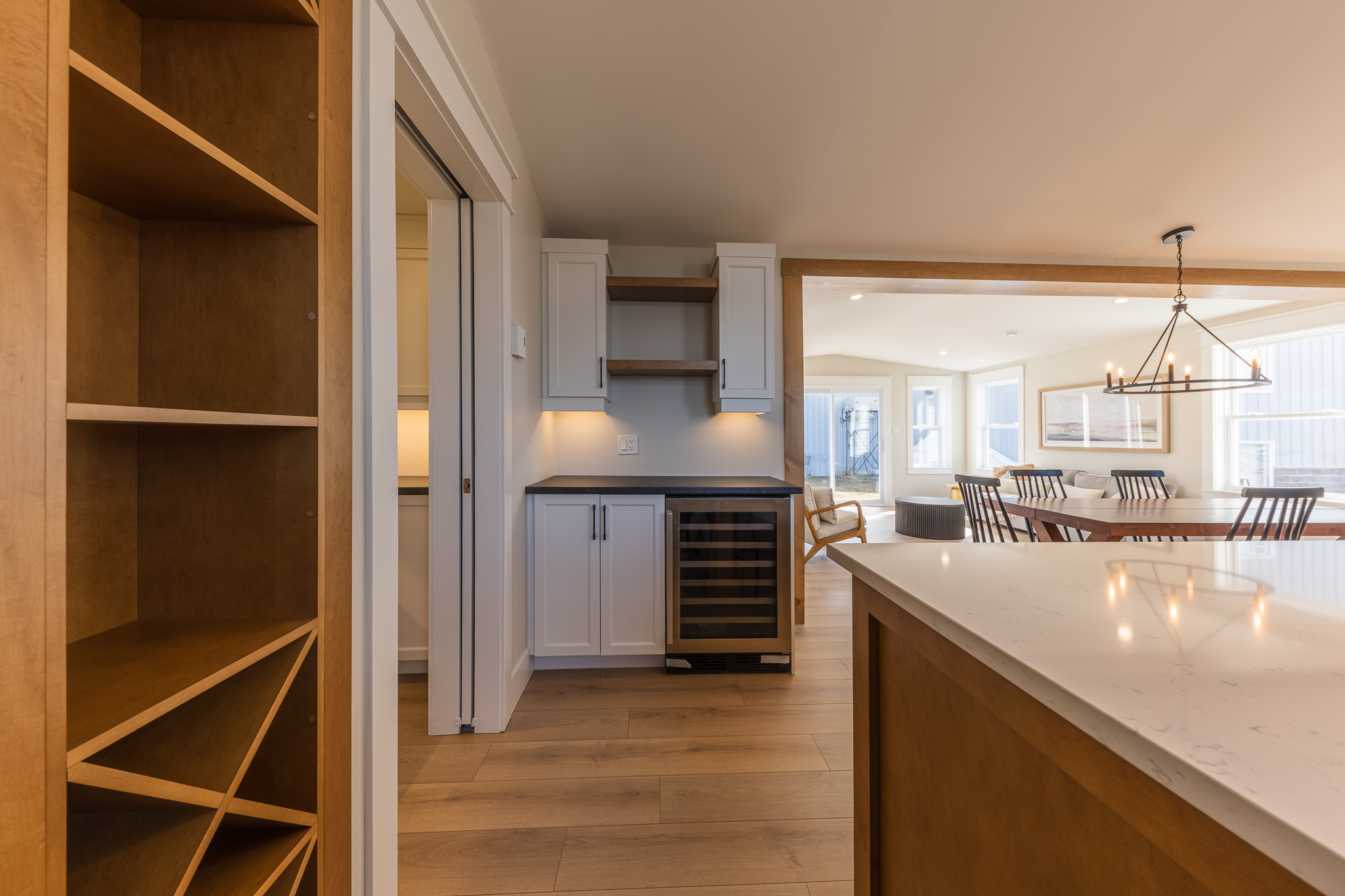 Modern kitchen and dining area with wooden floors. Features a wine fridge, white cabinets, shelving, a large dining table, and a black chandelier.