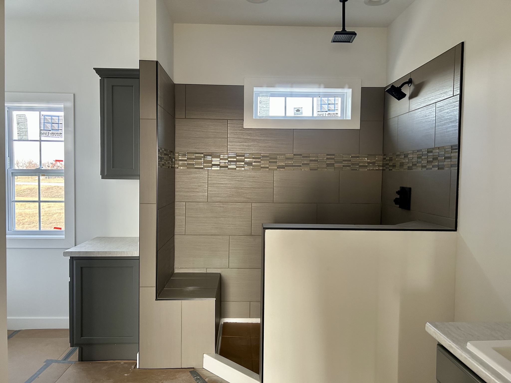 Modern bathroom with brown tiled walls and a mosaic accent strip. A window above provides natural light. Gray cabinets are seen to the left.