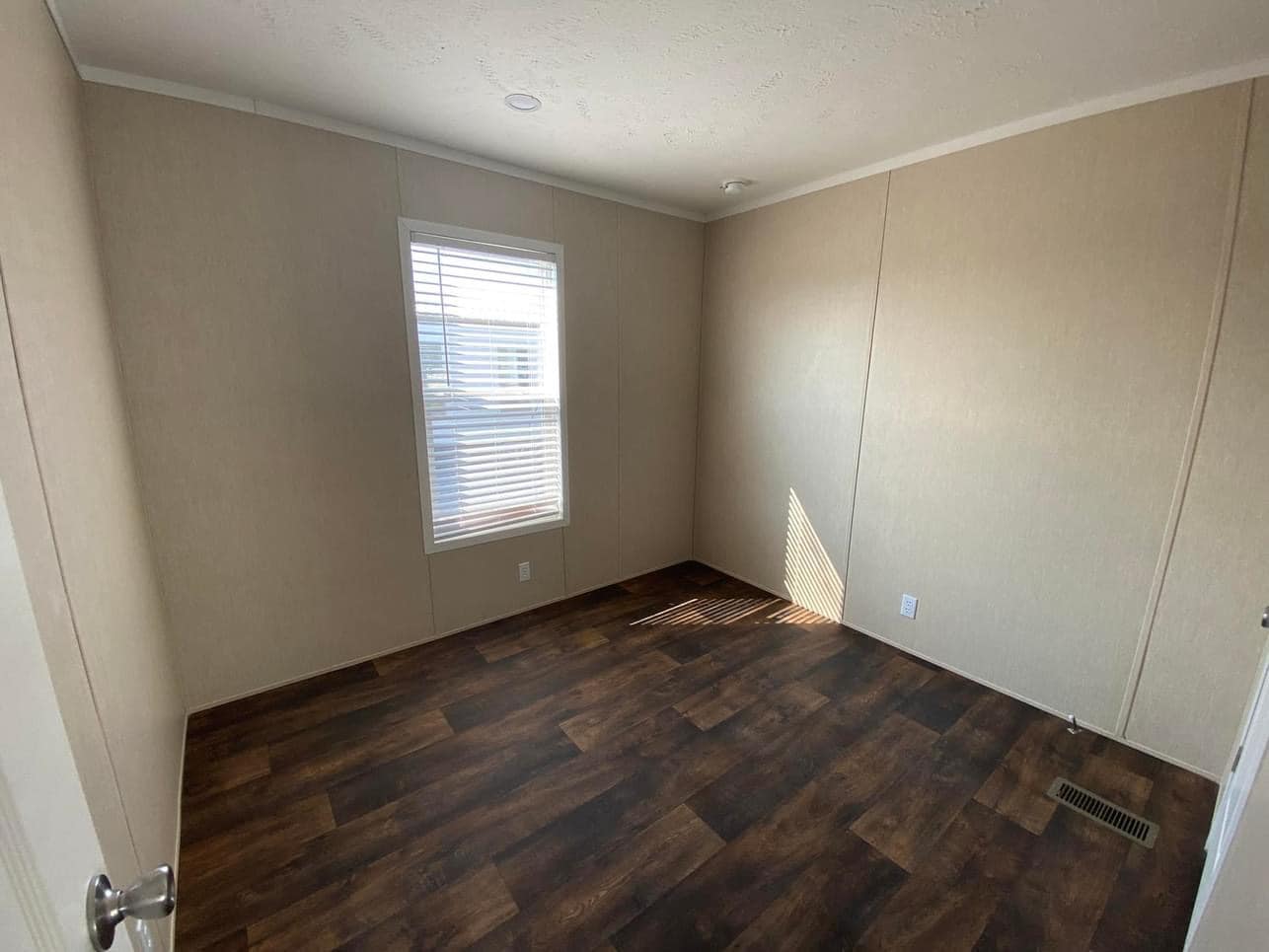 Empty room with beige walls and wood-style flooring. A single window with blinds casts soft shadows. The space appears clean and simple.