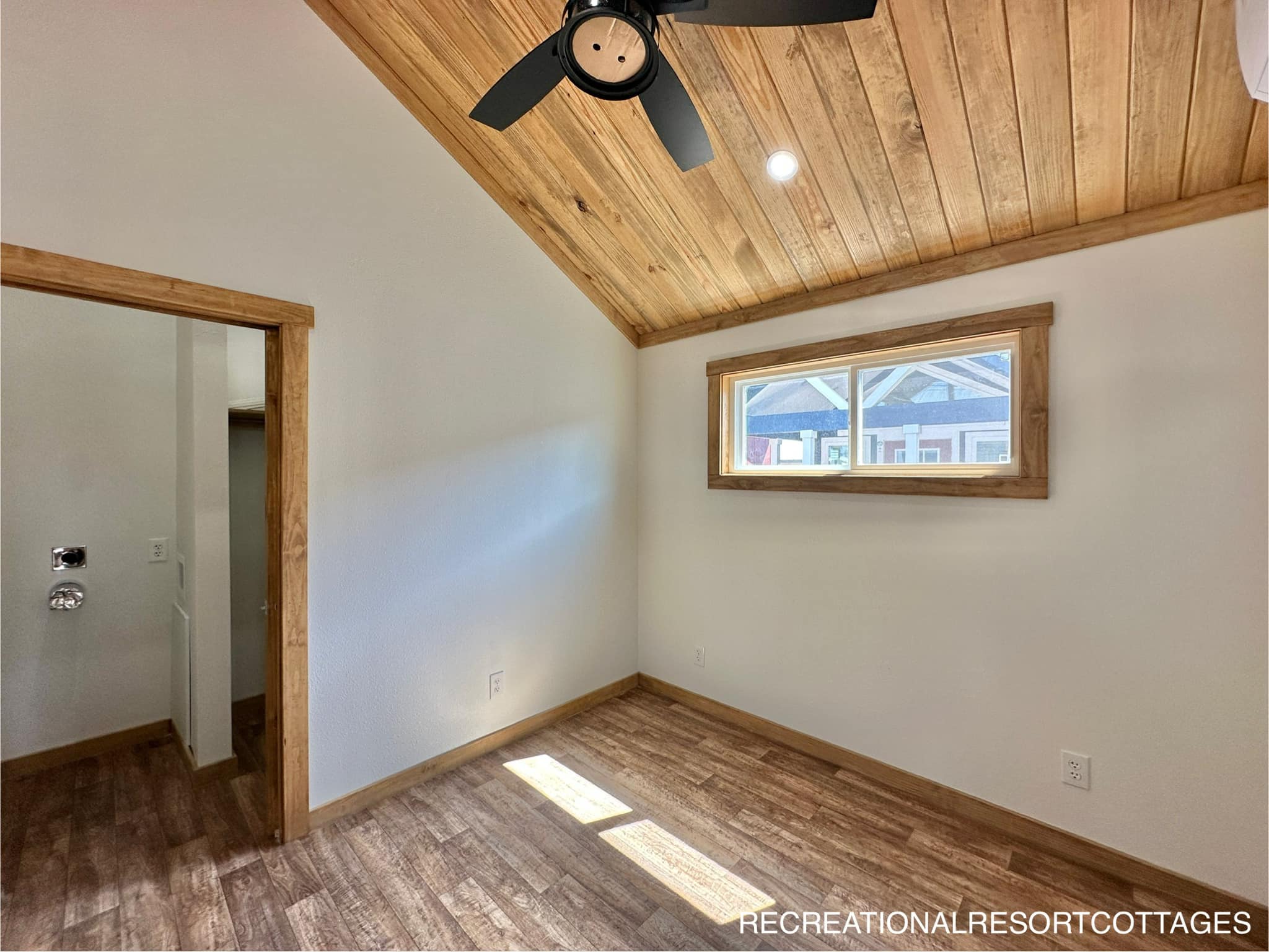 Cozy room with wood panel ceiling, white walls, and sunlight streaming through a rectangular window. A black ceiling fan adds a modern touch.