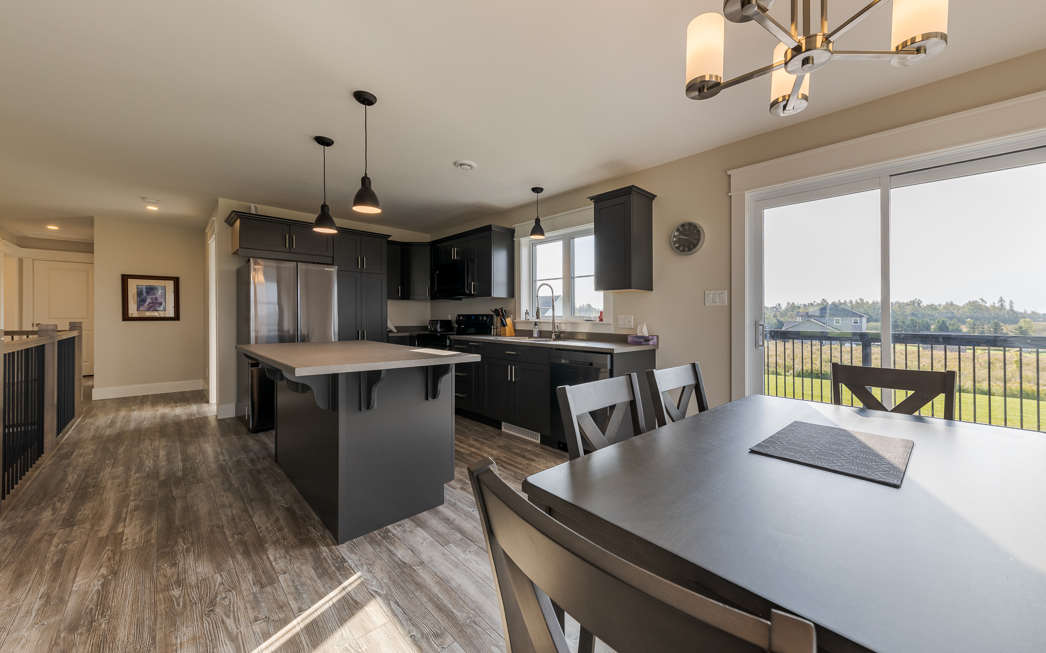 Modern kitchen and dining area featuring dark cabinets, a central island, and pendant lights. Large windows offer a view of a sunny backyard.