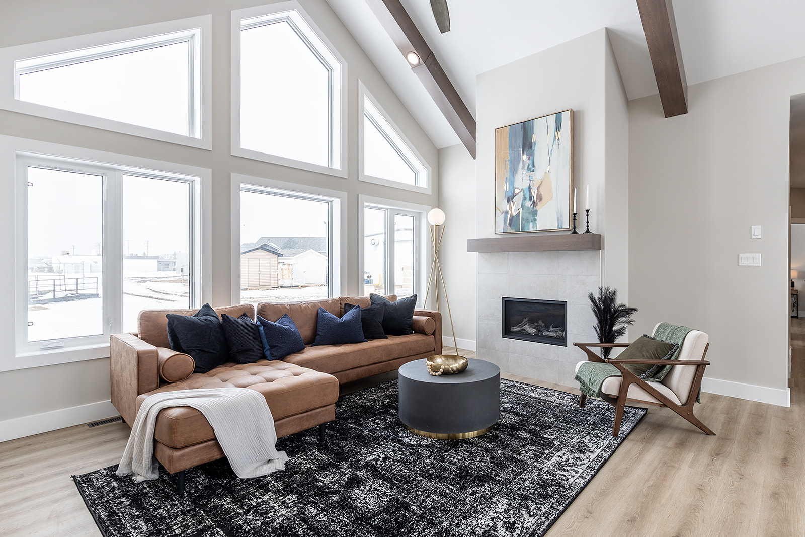 Modern living room with high ceilings, featuring a brown sectional sofa adorned with dark pillows, a patterned black rug, and a sleek fireplace. Large windows bring in natural light, creating an airy feel.