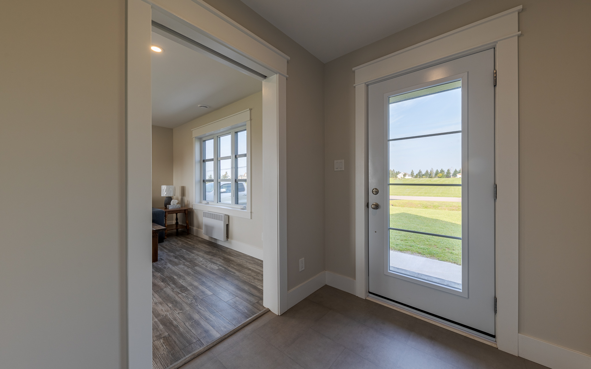 A bright room with wooden flooring seen through an open doorway. Adjacent, a glass door reveals a sunny, green landscape, creating a serene atmosphere.